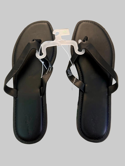 A pair of black leather thong sandals with flat soles, shown on a light-colored wooden floor. One sandal is on a clear plastic hanger, and the other has a paper tag attached.