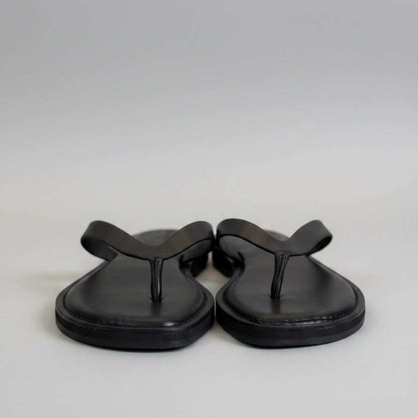 A pair of black leather flip-flop sandals with black straps and white plastic tags, resting on a light gray tabletop.