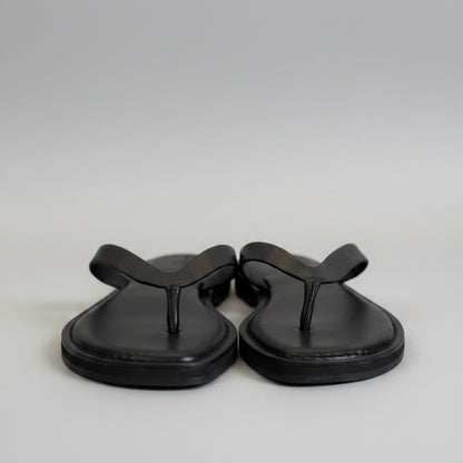 A pair of black leather flip-flop sandals with black straps and white plastic tags, resting on a light gray tabletop.