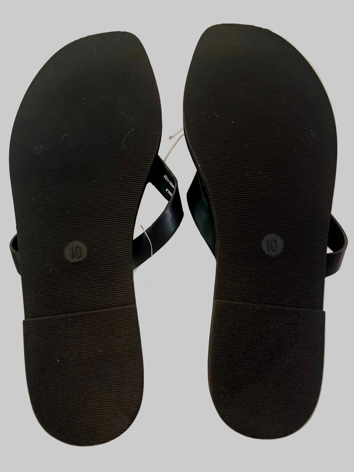 A pair of black flip-flop sandals shown from the bottom, displaying the textured black foam soles and thin black straps with a white size tag attached, set against a light-colored wooden floor.