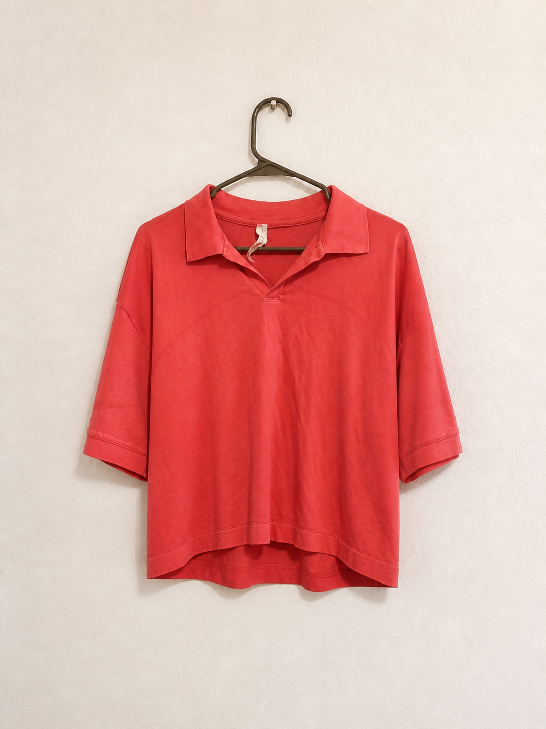 A coral red short-sleeve polo shirt hanging on a dark metal hanger against a light beige textured wall.