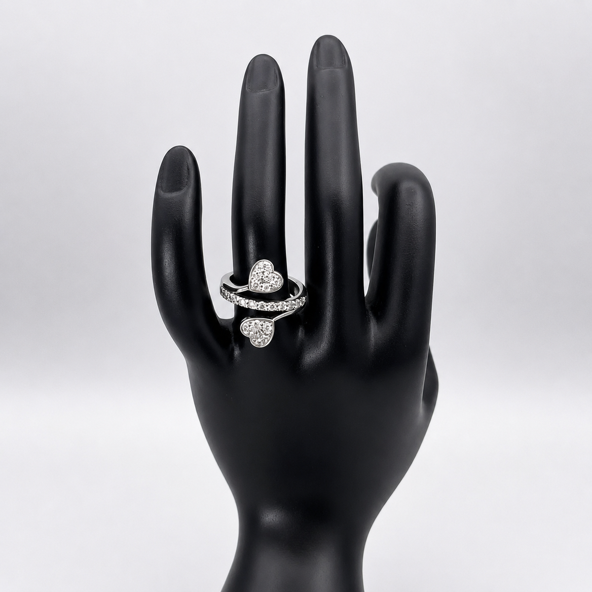 A silver ring with three heart-shaped pavé settings, displayed on a black mannequin hand against a light gray background.