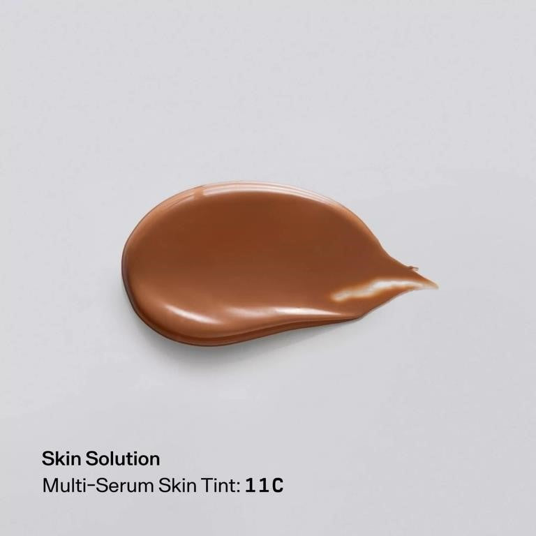 A teardrop-shaped smear of brown skin tint cosmetic product on a light gray background, with text below reading 'Skin Solution Multi-Serum Skin Tint: 11C'.