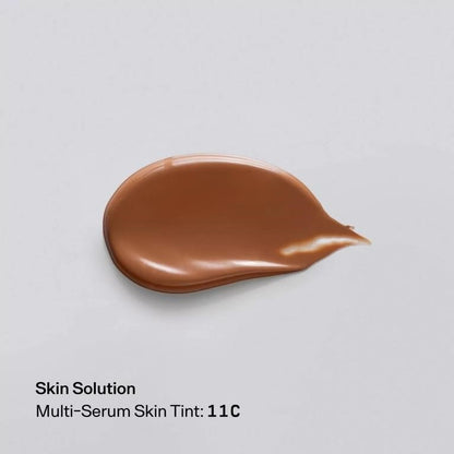 A teardrop-shaped smear of brown skin tint cosmetic product on a light gray background, with text below reading 'Skin Solution Multi-Serum Skin Tint: 11C'.