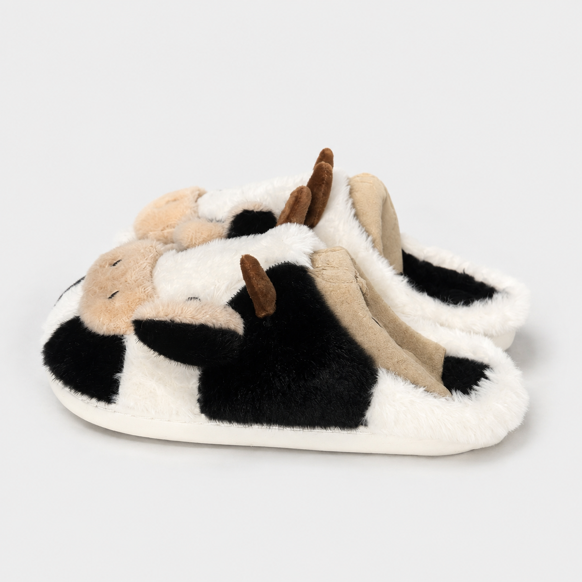 A pair of plush cow-themed slippers with black and white patches, beige face and belly details, and brown horns, shown on a light gray background.