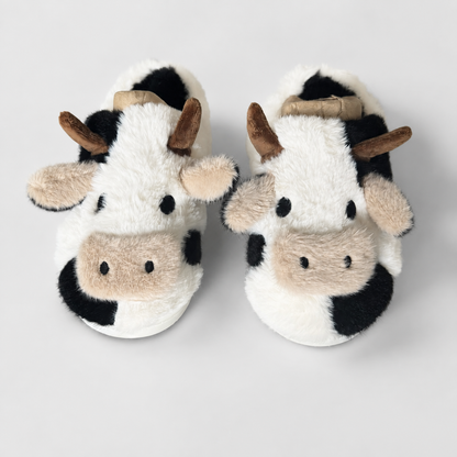 A pair of plush cow-themed slippers with black and white fur-like patterning, tan snouts and ears, and brown horns, photographed on a light gray background.