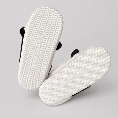 A pair of white slippers with black fuzzy trim and black bear ears, shown from the bottom to display the textured herringbone sole, on a light gray background.