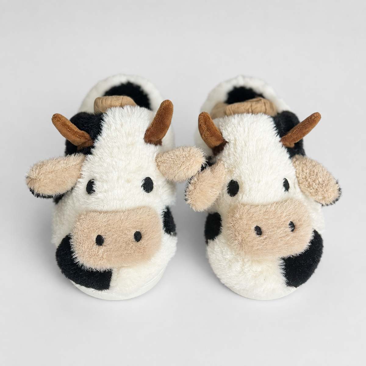 A pair of plush cow-themed baby slippers with white, black, and beige fur-like textures, brown horns, and embroidered eyes and nose details, shown on a light gray background.