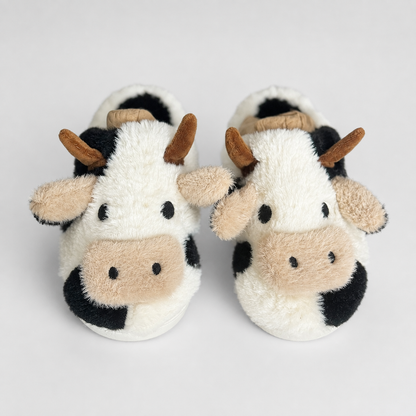 A pair of plush cow-themed baby slippers with white, black, and beige fur-like textures, brown horns, and embroidered eyes and nose details, shown on a light gray background.