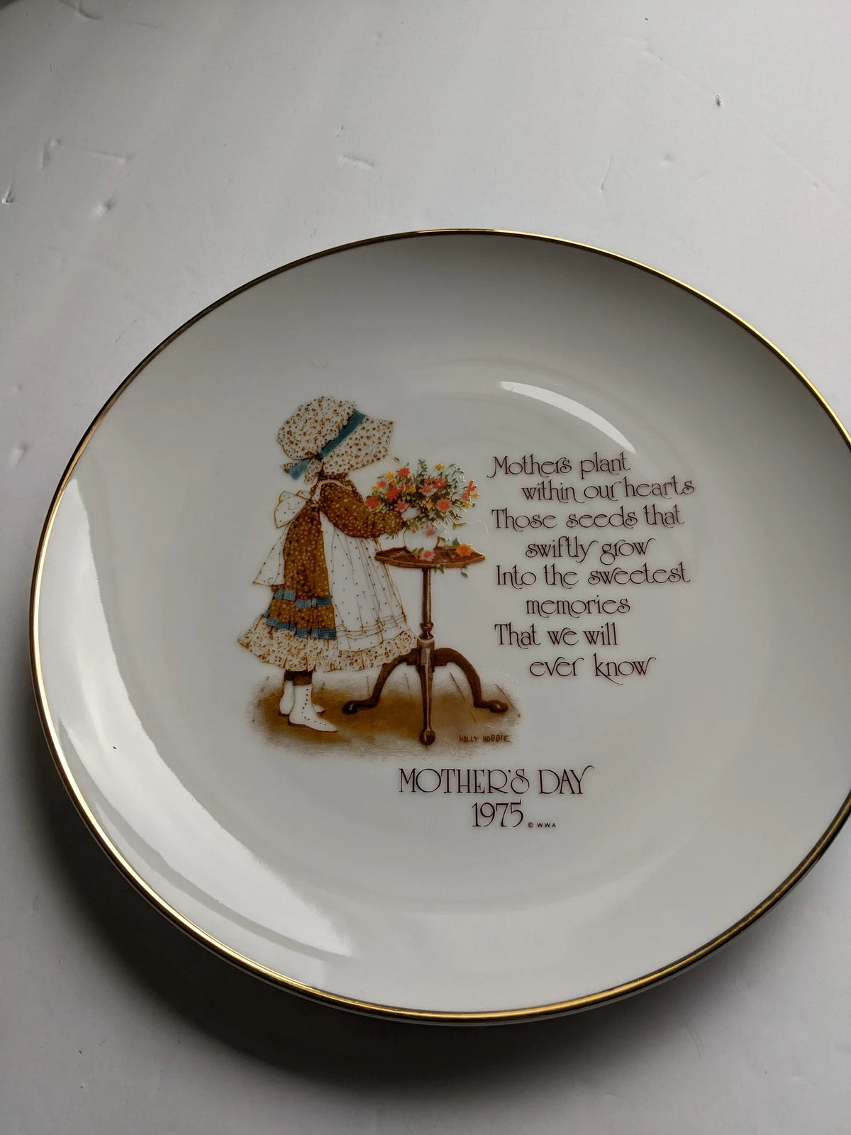 A white ceramic Mother's Day decorative plate with gold trim, featuring an illustration of a young girl watering flowers and a printed poem with the text 'MOTHER'S DAY 1975.