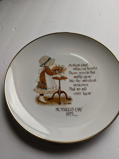 A white ceramic Mother's Day decorative plate with gold trim, featuring an illustration of a young girl watering flowers and a printed poem with the text 'MOTHER'S DAY 1975.