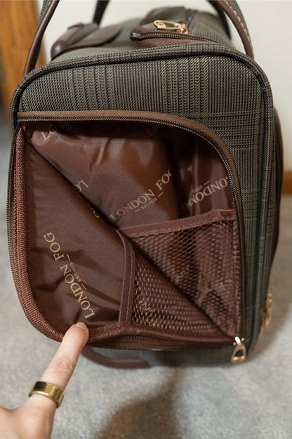 A close-up of a brown and black plaid London Fog suitcase or backpack, opened to show a brown interior organizer pocket with the brand name 'LONDON FOG' printed inside. A hand with a gold ring points toward the interior pocket.