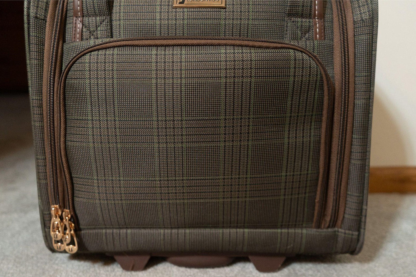 A close-up of a rolling suitcase showing its front pocket with a brown and olive green plaid pattern, gold-tone zipper pulls, and dark brown wheels on a carpeted floor.