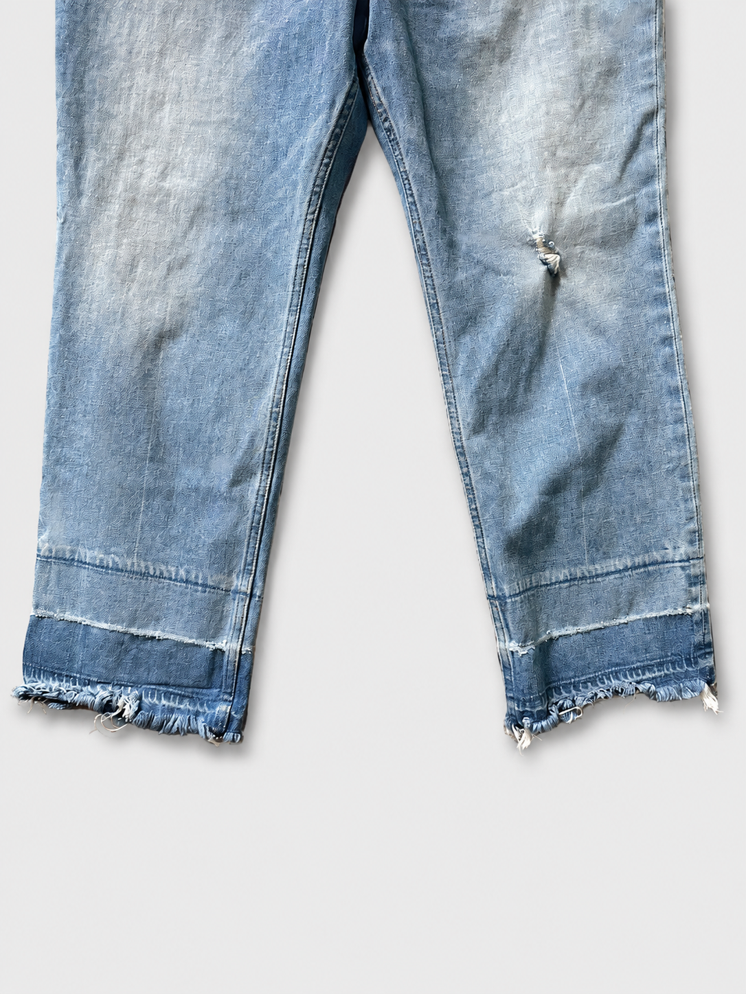 Close-up of light blue denim jeans with a raw, frayed hem and a small distressed rip on the right thigh, shown against a light gray background.