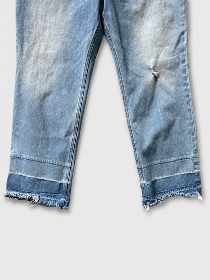 Close-up of light blue denim jeans with a raw, frayed hem and a small distressed rip on the right thigh, shown against a light gray background.