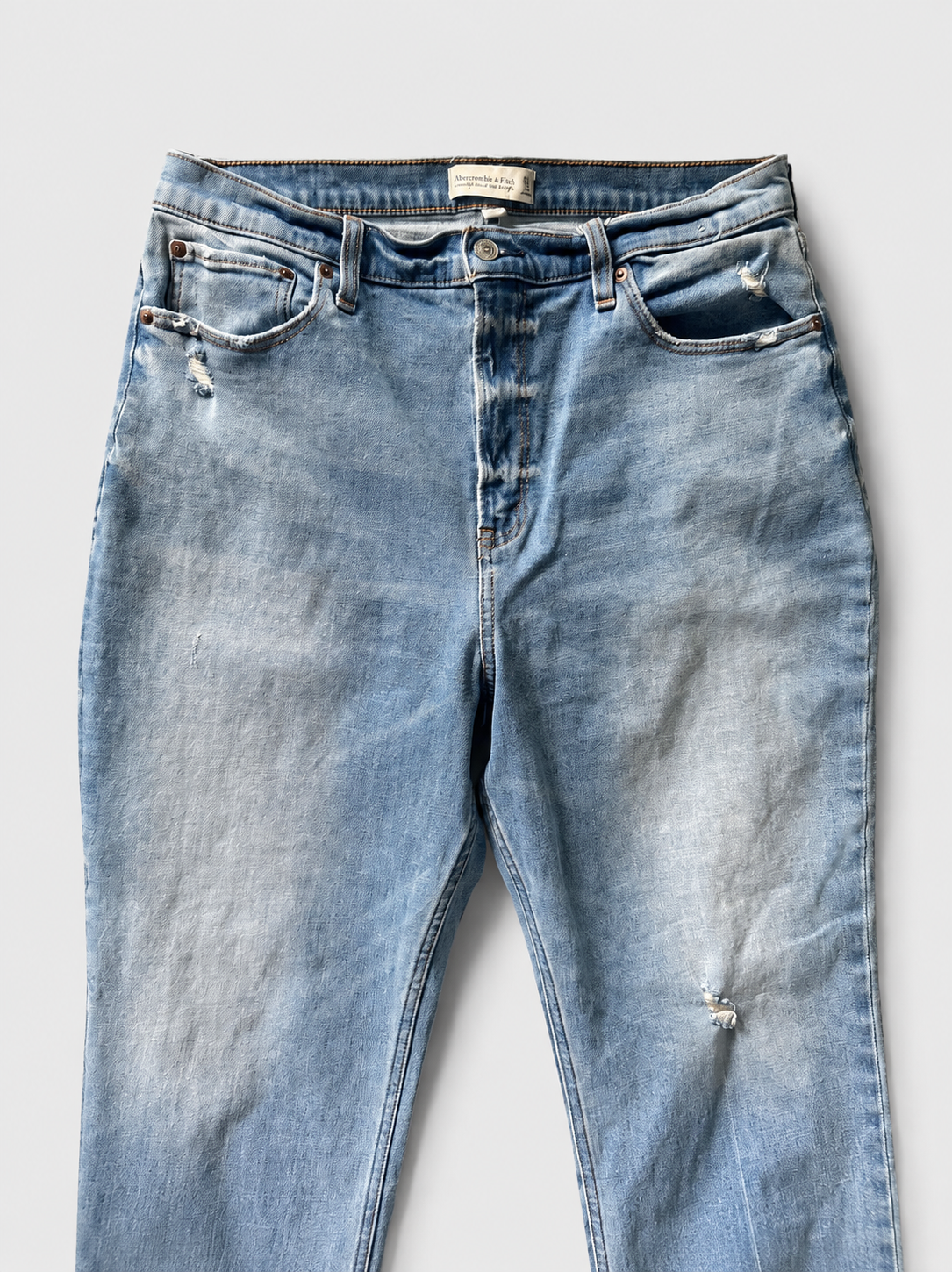 Light blue distressed denim jeans laid flat against a light gray background, featuring whiskering, fading, and small rips on the thighs.