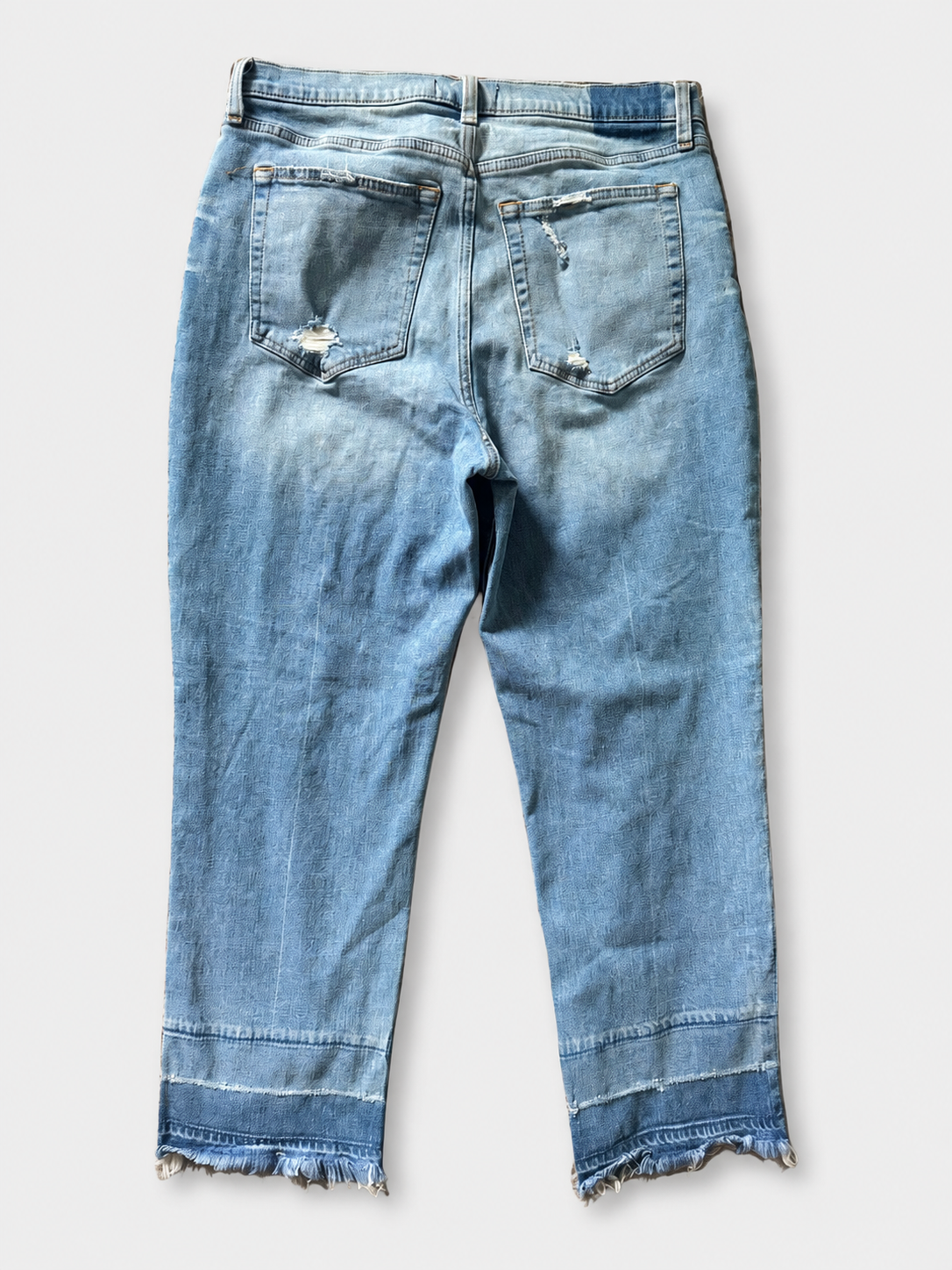 Light blue denim jeans shown from the back, featuring a raw frayed hem, distressed details on the back pockets, and a button closure, photographed against a light gray background.