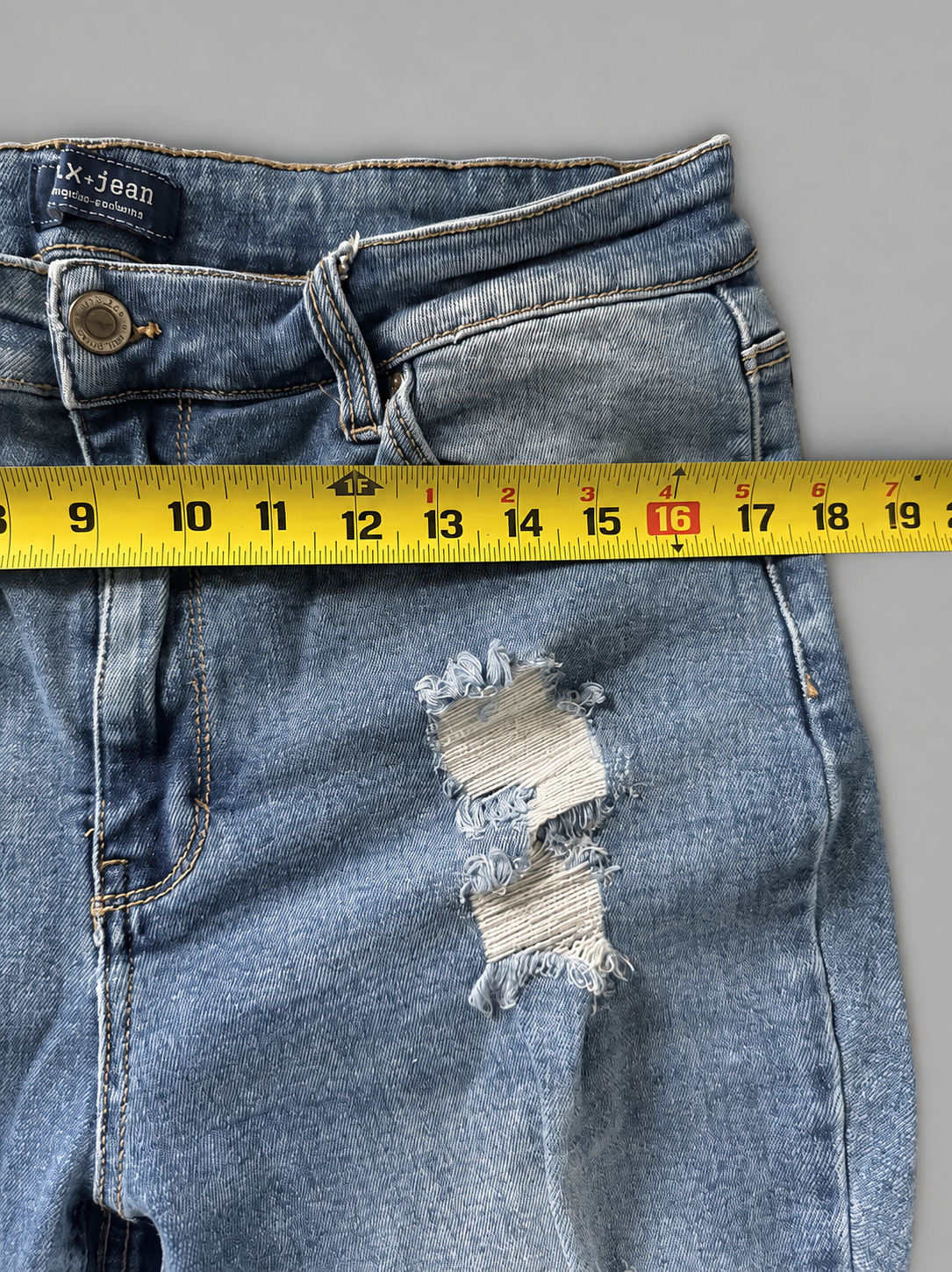 Close-up of a pair of light blue distressed denim jeans laid flat on a gray background, with a yellow measuring tape horizontally positioned across the thigh to show the fabric width.
