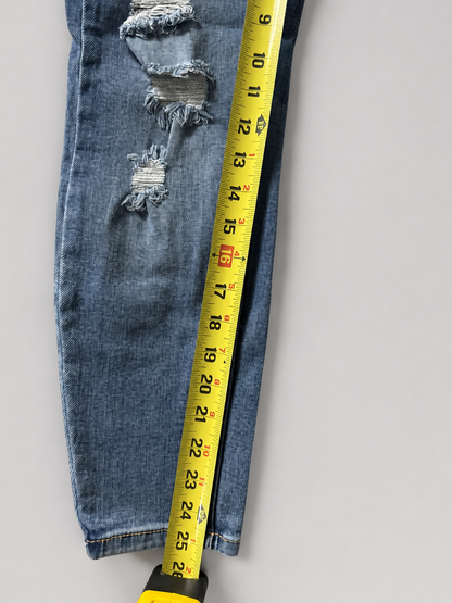 A close-up of a pair of blue denim jeans laid flat on a light gray background, with a yellow tape measure placed vertically along the side to show the leg length. The jeans have a distressed rip at the lower front.
