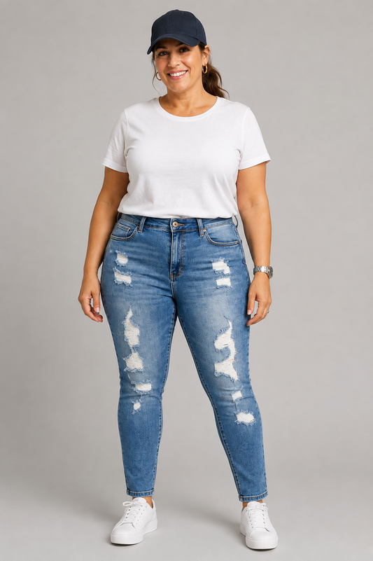A woman modeling a pair of high-waisted, distressed blue skinny jeans with a white t-shirt, white sneakers, and a navy baseball cap against a solid gray studio background.