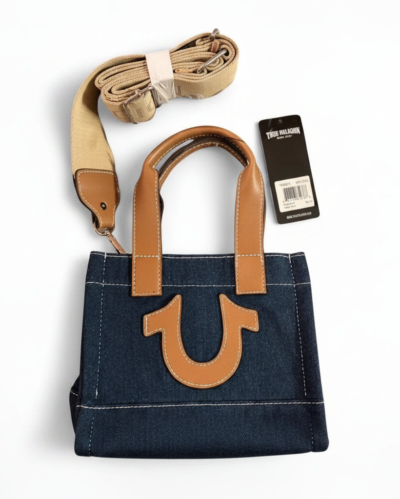 A denim tote bag with tan leather handles and a large tan U-shaped logo on the front, shown with a matching tan shoulder strap and a black product tag on a white background.