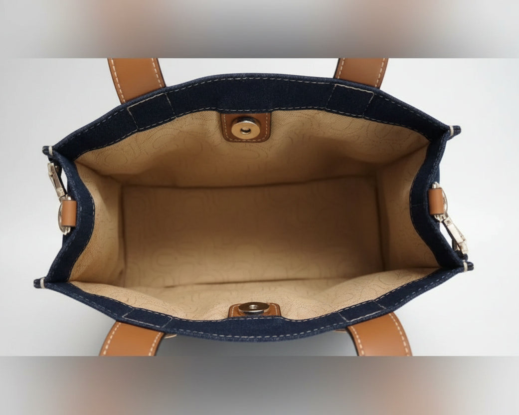 Close-up of the interior of a handbag with dark blue denim trim, tan leather trim, and a beige fabric lining, held open by a person's fingers.