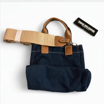 A dark blue denim tote bag with tan leather handles and trim, featuring a matching tan canvas shoulder strap with 'ERBLIG' text, shown on a white background with a black brand tag.