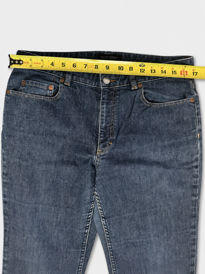 A pair of blue denim jeans laid flat on a light gray background, with a yellow tape measure stretched across the waistband to show the measurement.