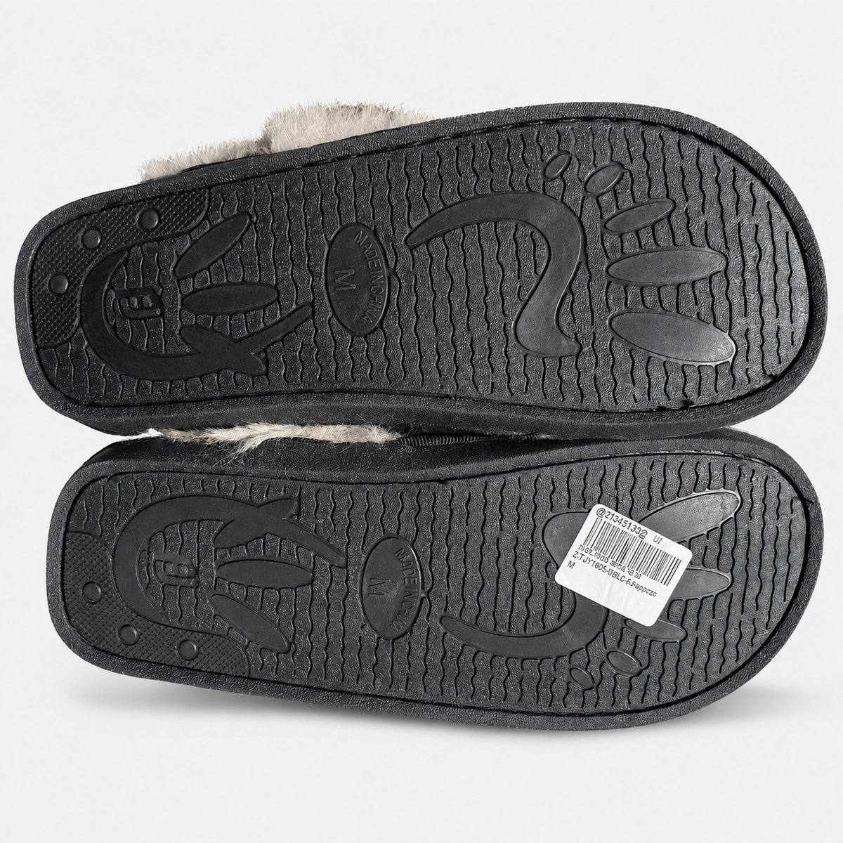 A pair of black slippers shown from the sole side, featuring a textured black rubber outsole with embossed footprints and a light beige faux-fur lining visible at the top edge. A white barcode sticker is on the bottom slipper.