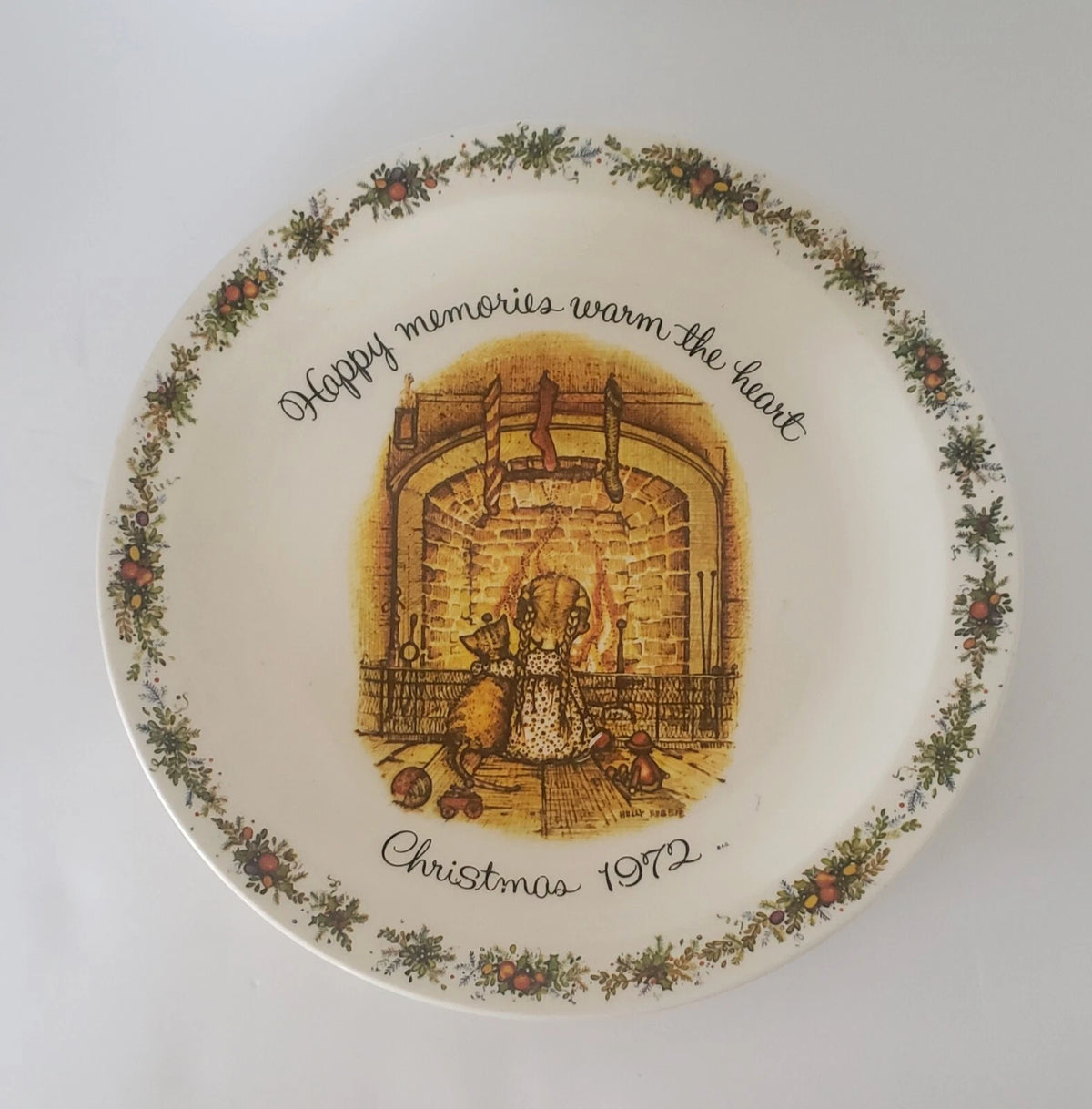 A round ceramic Christmas plate with a floral border and an illustration of a child by a fireplace. The plate features the text 'Happy memories warm the heart' and 'Christmas 1972'.