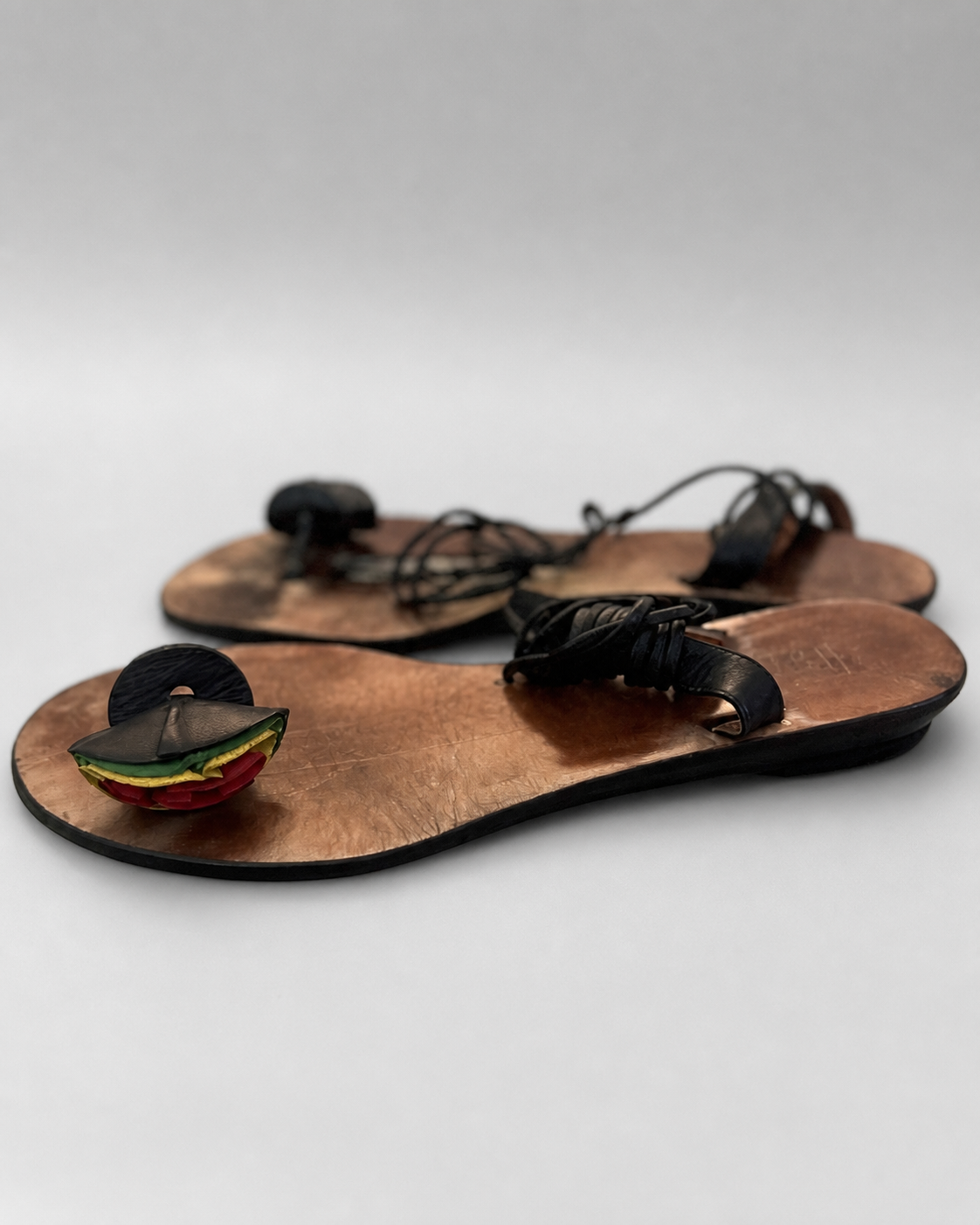A pair of handmade leather sandals with a copper-toned footbed, black woven straps, and a colorful decorative element on the front featuring red, yellow, and green layers.