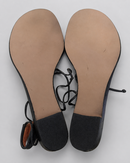 A pair of women's flat sandals shown from the bottom, featuring tan insoles, black suede-like heels, and thin black lace-up straps, photographed on a light gray background.