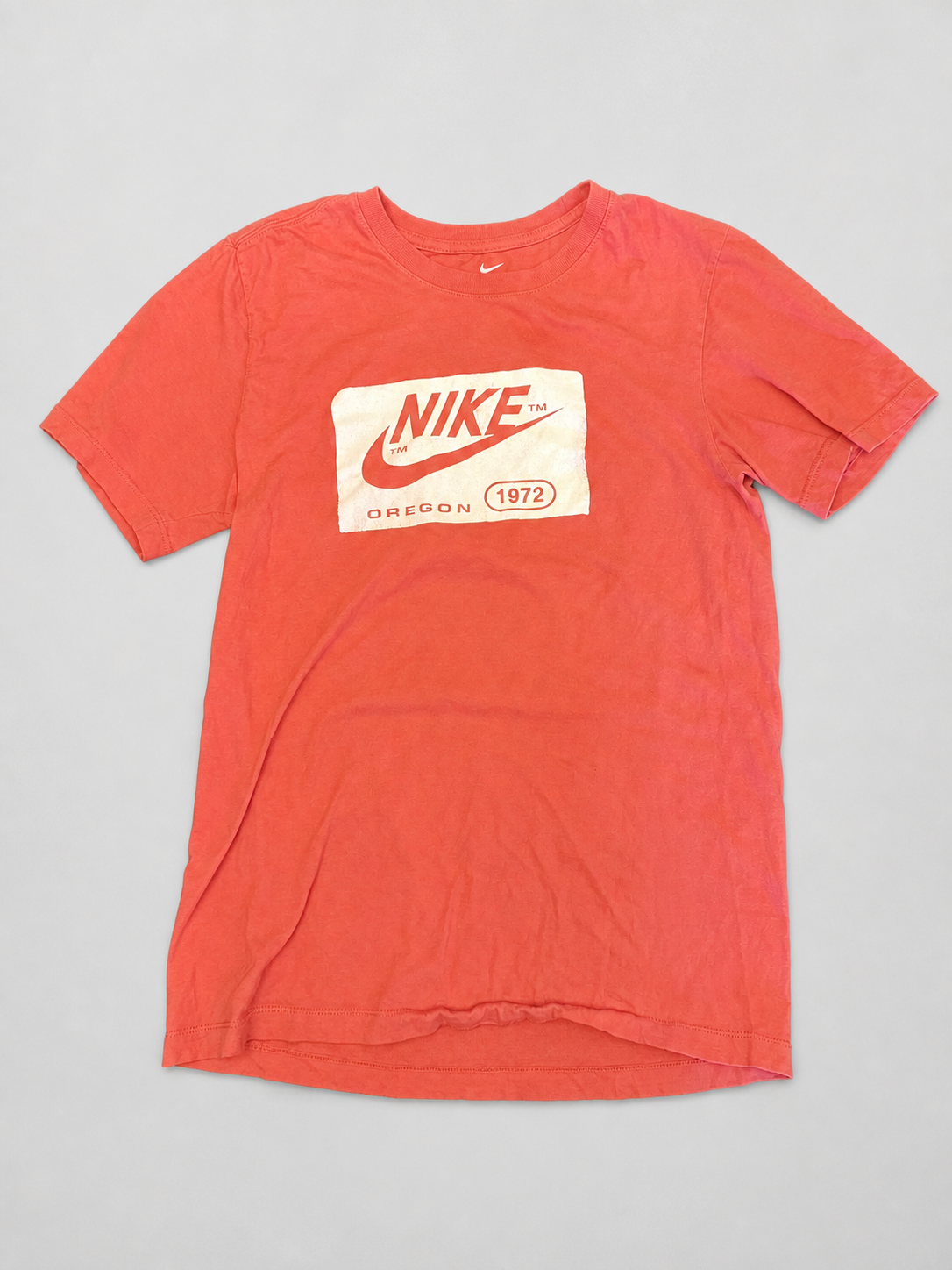 A faded red Nike short-sleeve t-shirt with a vintage-style cream graphic on the chest featuring the red swoosh and 'NIKE OREGON 1972' text, laid flat on a light gray background.