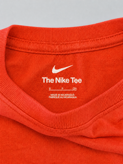 Close-up of the inside collar of a bright orange-red Nike t-shirt, showing the white printed Nike swoosh and 'The Nike Tee' text on the inner neck label.