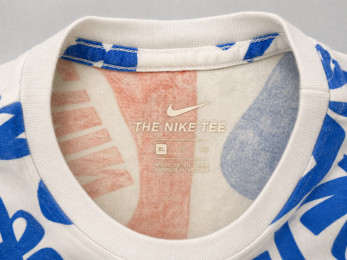 Close-up of the inside collar of a Nike t-shirt, showing the printed inner label that reads 'THE NIKE TEE' with a white Nike swoosh. The shirt features a white ribbed collar and blue and pink abstract graphic patterns on the sleeves and body.