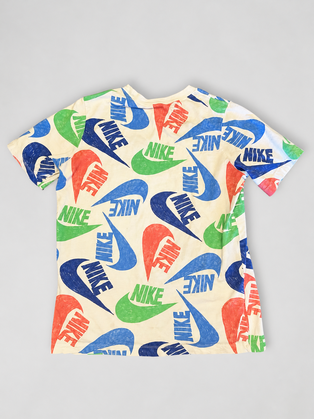 A short-sleeve crewneck t-shirt featuring an all-over print of the Nike Swoosh and 'NIKE' text in blue, green, and orange-red on a cream-colored background, laid flat on a light gray surface.