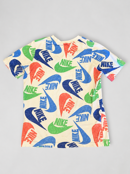 A short-sleeve crewneck t-shirt featuring an all-over print of the Nike Swoosh and 'NIKE' text in blue, green, and orange-red on a cream-colored background, laid flat on a light gray surface.