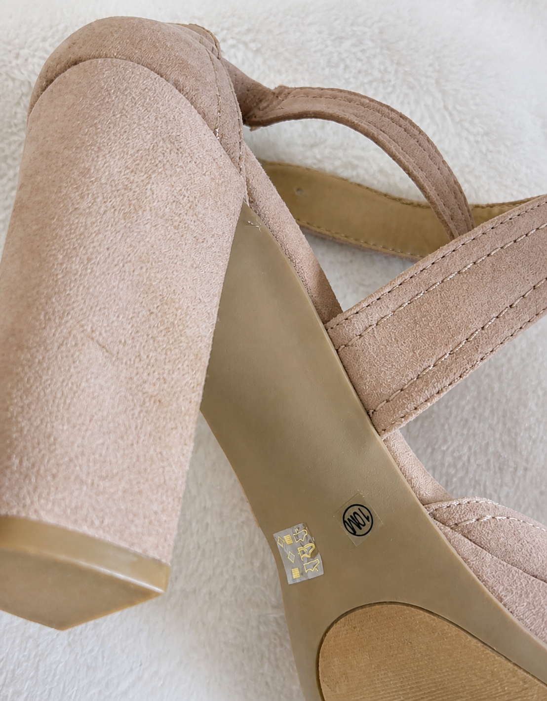 A close-up of a beige suede block-heel sandal with a low-cut toe and a small gold logo on the insole, resting on a white textured fabric background.