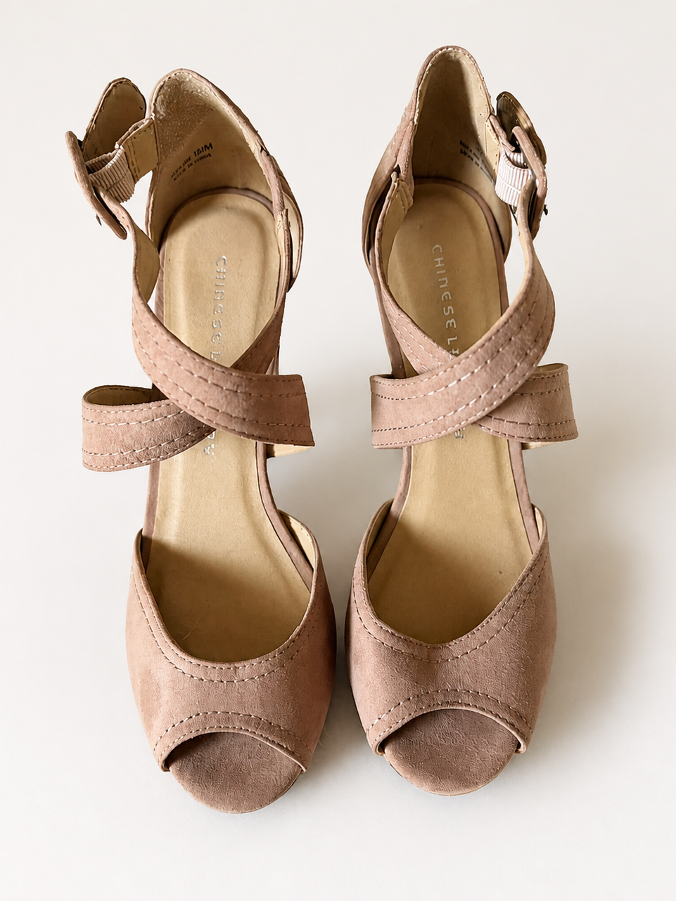 A pair of women's peep-toe block-heel sandals in a dusty taupe suede-like material, featuring crisscross ankle straps and side buckle closures, photographed from a top-down angle on a light off-white background.