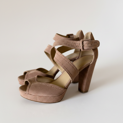 A pair of taupe brown suede high-heeled sandals with open toes, crisscross ankle straps, and block heels, photographed on a clean light gray studio background.